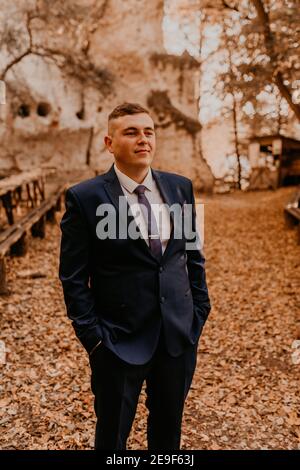 Ein Mann in einem formalen Anzug der Bräutigam steht im Herbstwald hält er seine Hände in seinen Taschen. Rock Kloster in Bakota Stockfoto