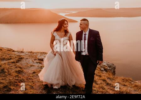 Liebevolles Paar Hochzeit Brautpaar in einem weißen Kleid und Anzug Spaziergang im Sommer auf dem Berg über dem Fluss. Sonnenuntergang und Sonnenaufgang. Mann und Frau auf dem Stockfoto