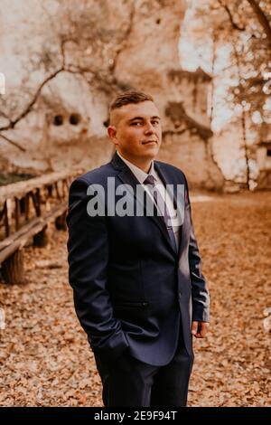 Ein Mann in einem formalen Anzug der Bräutigam steht im Herbstwald hält er seine Hände in seinen Taschen. Rock Kloster in Bakota Stockfoto