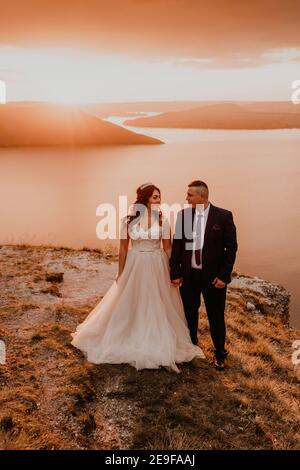 Liebevolles Paar Hochzeit Brautpaar in einem weißen Kleid und Anzug Spaziergang im Sommer auf dem Berg über dem Fluss. Sonnenuntergang und Sonnenaufgang. Mann und Frau auf dem Stockfoto