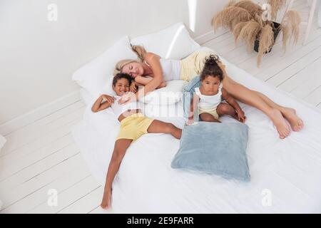 Junge kaukasische blonde Mutter spielt, liegt auf weißem Bett mit afroamerikanischen Tochter Sohn. Happy multi ethnischen Familie Morgen. Frau umarmt, streichelt Kind Stockfoto