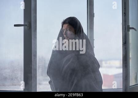 Kaltes Winterwetter. Eine Frau zittert aus der Kälte, die auf der Fensterbank sitzt, eingewickelt in eine Decke. Frostige Luft strömt aus dem offenen Fenster hinein. Airi Stockfoto