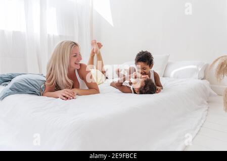Junge kaukasische blonde Mutter spielt, liegt auf weißem Bett mit afroamerikanischen Tochter Sohn. Happy multi ethnischen Familie Morgen. Frau umarmt, streichelt Kind Stockfoto