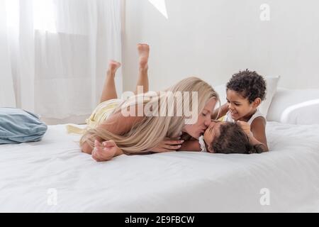 Junge kaukasische blonde Mutter spielt, liegt auf weißem Bett mit afroamerikanischen Tochter Sohn. Happy multi ethnischen Familie Morgen. Frau umarmt, streichelt Kind Stockfoto