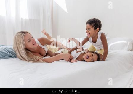 Junge kaukasische blonde Mutter spielt, liegt auf weißem Bett mit afroamerikanischen Tochter Sohn. Happy multi ethnischen Familie Morgen. Frau umarmt, streichelt Kind Stockfoto