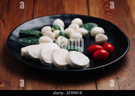 Weiße kleine Mozzarella Käsebällchen, Spinatblätter und Tomaten auf schwarzem Teller. Stockfoto