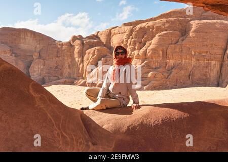 Junge Frau in warmer Jacke und Schal um den Kopf lächelnd, sitzt an natürlich geformten Felsenfenster in wadi Rum Wüste an sonnigen Tag Stockfoto