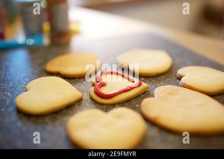 Nahaufnahme von herzförmigen Keksen auf einer Arbeitsfläche in der Küche. Ein Biskuit ist teilweise dekoriert, die anderen werden gerade dekoriert. Stockfoto