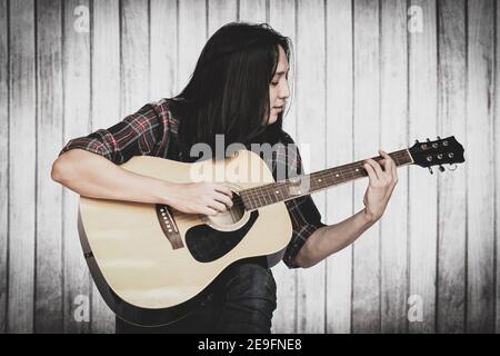 Portrait schöner junger männlicher Gitarrist mit langen Haaren sitzen und spielen akustische Gitarre. Asiatische langhaarige Mann spielen akustische Gitarre Holz Wand Bac Stockfoto