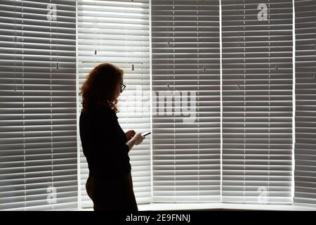 Foto einer jungen Frau in einer Shilouette, die während der Isolation des britischen Covid 19 vor einem großen Fenster stand. Frau hält ein Mobiltelefon. Stockfoto