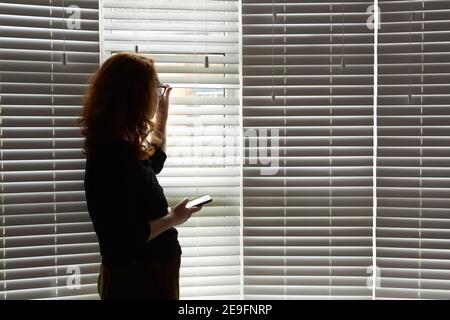 Foto einer jungen Frau in einer Shilouette, die während der Isolation des britischen Covid 19 vor einem großen Fenster stand. Frau hält ein Mobiltelefon. Stockfoto