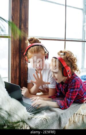 Zwei rothaarige Teenager-Mädchen in roten Kopfhörern hören fröhlich Musik, neben einem Laptop, sitzen auf dem Fenster Stockfoto