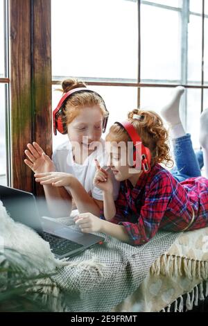 Zwei rothaarige Teenager-Mädchen in roten Kopfhörern hören fröhlich Musik, neben einem Laptop, sitzen auf dem Fenster Stockfoto