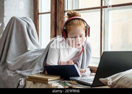 Ein Teenager-Mädchen mit roten lockigen Haaren genießt es, Musik auf dem Fensterbrett zu hören. Online-Training, hört sich Vorträge an Stockfoto