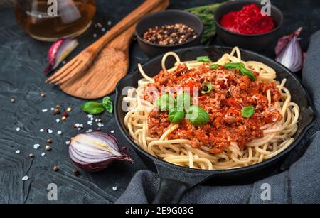 Spaghetti und Sauce in einer Pfanne werden mit Käse und frischem Basilikum auf einem Hintergrund von Gewürzen und Zwiebeln auf einem schwarzen Betongrund bestreut. Stockfoto