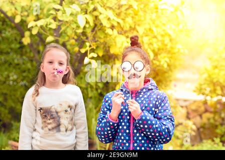 Überrascht kleines Mädchen hält lustige Lippen auf Stick und glückliches Mädchen mit niedlichen Sonnenbrillen und Noisemaker auf grüne Natur kopieren Raum Hintergrund. Purim, 1 April Narr Tag , Geburtstag Party Konzept Stockfoto