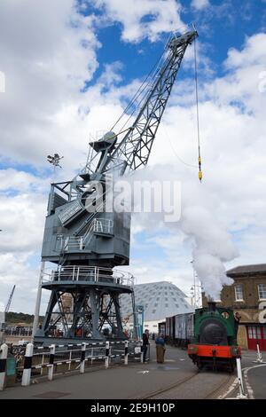 Großer alter Schiffsbau / Schiffbaukran mit der Lokomotive 'Ajax' aus dem Zweiten Weltkrieg, ein Dampflokomotiv, der darunter vorbeifährt, in Chatham Historic Dockyard, Kent England (121). Stockfoto