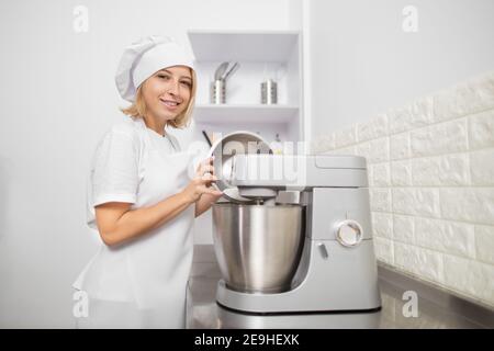 Attraktive Konditorei Mädchen in weißem Hut und Schürze, lächelnd an der Kamera in der modernen hellen Küche, die Vorbereitung eines Dessert, Kuchen oder Kekse, mit Planetar Stockfoto