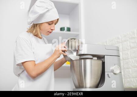 Junge blonde Mädchen, professionelle Konditorei, trägt weiße Schürze und Kochmütze, Kochen in gemütlichen hellen Küche Interieur, mit Mixer oder Küchenmaschine Stockfoto