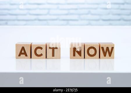 'Act Now'-Message-Schild auf Holzblöcken auf einem weißen Tisch mit weißen Brinks auf dem Hintergrund sitzen. Stockfoto