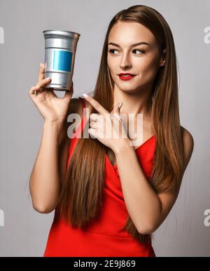 Junge Frau mit langen seidigen Haaren in roten Kleid zeigt Finger am Behälter mit Haarpflege Behandlung Kosmetik hält sie Stockfoto