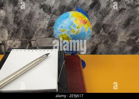 Auf dem Schreibtisch liegen ein großes Lexikonbuch und ein Notizbuch mit Stift. Artikel Buch und Brille, Globus und Notizbuch auf dem Tisch. Hintergrund mit Leerzeichen fo Stockfoto