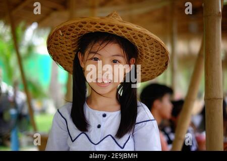 Ein süßes junges asiatisches Mädchen, das einen traditionellen Bambus gewebten Bauernhut auf einer Reisfarm in Thailand trägt und sich glücklich und lächelnd fühlt. Stockfoto