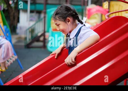 Ein nettes junges asiatisches Mädchen in Schuluniform rutscht eine rote Plastikrutsche auf einem Schulhof während der Aussparung hinunter. Stockfoto