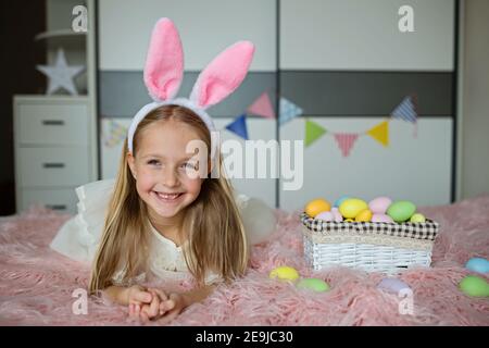 Glücklich kaukasischen Kleinkind Mädchen 7 Jahre alt trägt Hasenohren Stirnband, liegen auf dem Bett zu Hause im Schlafzimmer mit farbigen ostereiern. Bleiben Sie zu Hause während Stockfoto