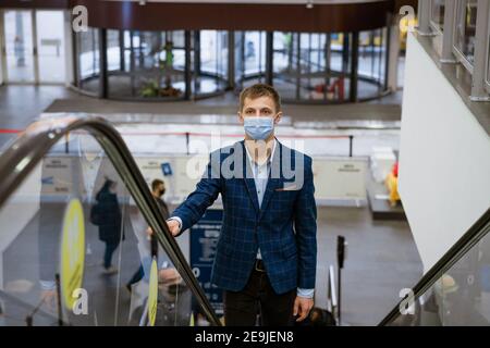 Junger Kaukasier in einer Jacke und einer medizinischen Maske Steigt die Rolltreppe in einem Einkaufszentrum Stockfoto