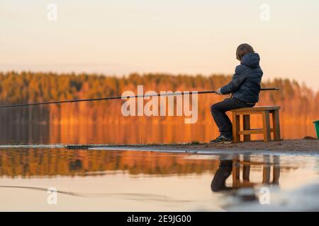 Angeln am Abend. Kleiner Junge mit Angelrute in den Händen auf dem See. Betrachtet den hellen Herbstwald, der sich im Wasser spiegelt. Stockfoto