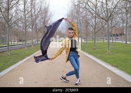 Hübscher junger Mann, der Make-up und Kopfhörer trägt und mit einem Baumwollschal in einem Parkpfad tanzt. Nicht binäre androgyne Kerl. Stockfoto