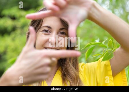 Nahaufnahme einer lächelnden jungen Frau, die ihr Gesicht mit ihren Händen als Selbstporträt umrahmt. Sie ist in der Natur. Leerzeichen für Text. Stockfoto