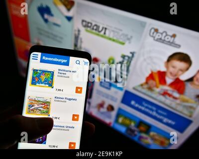 Person mit Smartphone mit Online-Shop des deutschen Spiel- und Spielzeugunternehmens Ravensburger AG auf dem Display vor der Website. Fokus auf Handy-Bildschirm. Stockfoto