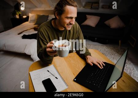 Kaukasischer männlicher Student genießen heißen Kaffee, während Sie sich online anmelden Unterricht auf Laptop sitzen am Schreibtisch im Home Office Stockfoto