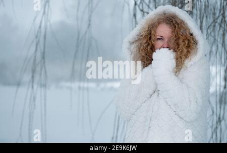 Rotschopf reife Frau in ihren fünfziger Jahren mit roten lockigen Haaren Mit weißem Kapuzenmantel in der Schnee-Winterlandschaft Stockfoto