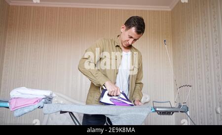 Konzentrierter Mann trägt braunes Hemd tut Hausarbeiten und bügelt Kleidung Nach der Wäsche mit lila Bügeleisen auf spezielle Bord schließen Anzeigen Stockfoto