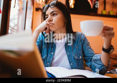 Erschöpfte Studentin sitzt am Tisch im Café und hält eine Tasse Kaffee und schaut aus dem Fenster. Stockfoto