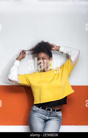 Positive afroamerikanische Millennial weiblich in trendigen gelben Shirt berühren Lockiges Haar und wegschauen, während man in der Nähe der Wand tanzt Stockfoto