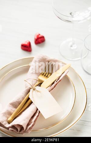 Romantische Tischkulisse mit goldenen Rim-Tellern, Bonbon-Herzen und goldenem Besteck mit Anhänger auf weißem Tisch. Stockfoto