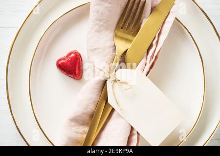 Romantische Tischkulisse mit goldenen Rim-Tellern, Bonbon-Herz und goldenem Besteck mit Anhänger auf weißem Tisch. Nahaufnahme, Idee für Valentinstag. Stockfoto