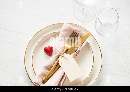 Romantische Tischkulisse mit goldenem Rand Teller, Bonbon Herz, Gläser und goldenes Besteck mit Anhänger auf weißem Tisch. Draufsicht. Stockfoto