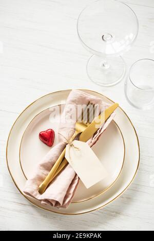 Romantische Tischkulisse mit goldenem Rand Teller, Bonbon Herz, Gläser und goldenes Besteck mit Anhänger auf weißem Tisch. Stockfoto