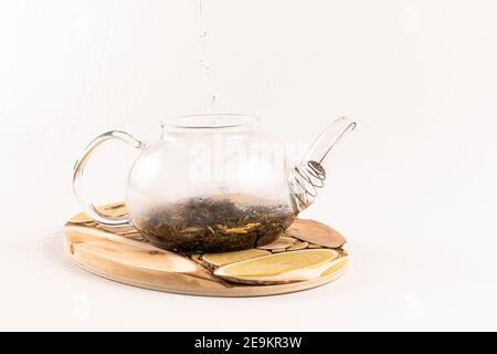 Kochendes Wasser wird in eine Glas Teekanne mit aromatischen gegossen Kräutertee steht auf einem hölzernen Wacholderständer auf einem Weißer Hintergrund Stockfoto