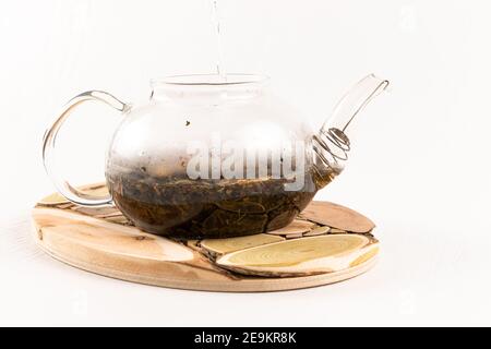 Kochendes Wasser wird in eine Glas Teekanne mit aromatischen gegossen Kräutertee steht auf einem hölzernen Wacholderständer auf einem Weißer Hintergrund Stockfoto