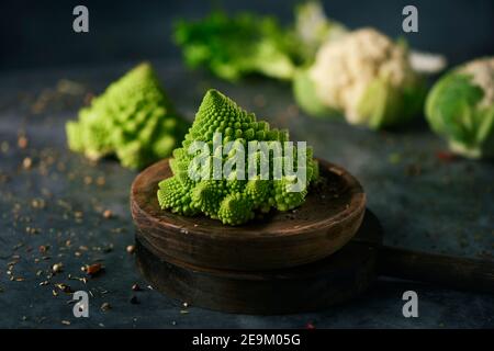 Ein Baby romanesco Brokkoli Kopf auf einem Holzteller, platziert auf einer dunklen Steinoberfläche neben einem anderen Baby romanesco Brokkoli Kopf und einige Baby cauli Stockfoto