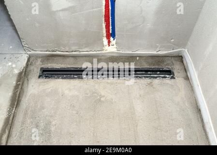Ein moderner schwarzer linearer Duschabfluss im Duschboden, sichtbare Wasserleitungen in der Wand. Stockfoto