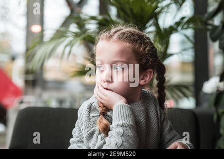 Hübsches junges Mädchen mit Zöpfen grau wollig Pullover saß in Moderne Schulumgebung mit Blick aus dem Fenster natürliches Licht Tag träumen Gelangweilter Kopf in den Händen Stockfoto