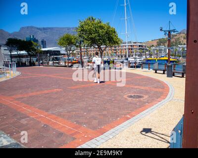 Waterfront - Kapstadt, Südafrika - 03-02-2021 Ziegelsteinpflasterter Gehweg, der zum Silo District führt. Mann, der im Vordergrund vorbei geht. Hafen und Tafelberg. Stockfoto