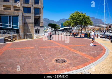 Waterfront - Kapstadt, Südafrika - 03-02-2021 Mann zu Fuß und Mann Motorrad im Vordergrund. Hafen und Tafelberg im Hintergrund. Stockfoto